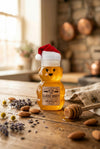 Honey bear bottle with a Santa hat on a wooden surface with honey, almonds, and lavender.