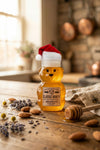 Honey bear bottle with a Santa hat on a wooden surface with honey, almonds, and lavender.