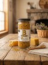 Jar of B Pollen on a wooden table with honeycomb and wooden spoon in a warm, homey setting.