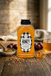 Bottle of Big Bear Honey on a wooden table with honeycomb pieces and plums.