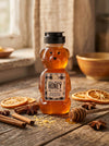 Bear shaped honey bottle on wooden table with honey dipper and spices surrounding it.