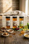 Three bottles of honey on a wooden table with honeycomb, honey dipper, and fruits.