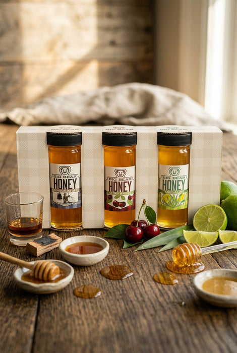 Three bottles of honey on a wooden table with honeycomb, honey dipper, and fruits.
