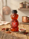 Honey bear bottle labeled 'Hot' on a wooden surface with spices and a bowl.