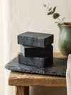 Three black soap bars stacked on a slate tray with a vase in the background.