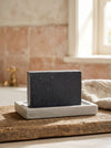 Black soap bar on a white plate with a bathroom setting in the background