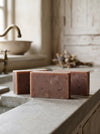 Three brown soap bars on a stone surface with a bathroom setting in the background.