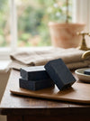 Three black soap bars on a wooden surface with a blurred natural background