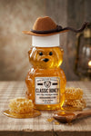 Honey bear with a cowboy hat on a wooden table with honeycomb and a spoon.